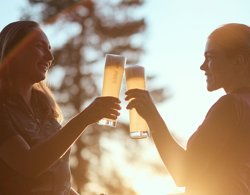 Zwei Frauen stehen auf einer Terrasse. Sie stoßen mit einem Bier an.