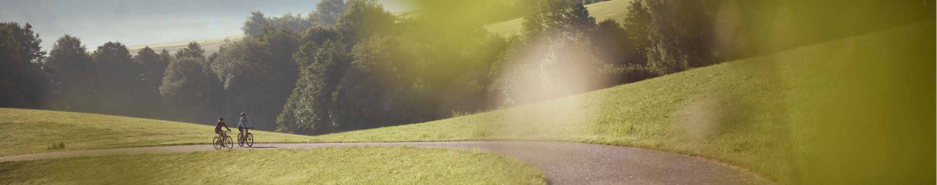 Zwei Frauen fahren mit dem Fahrrad bergab auf einer asphaltierten Straße. Im Hintergrund ist eine hügelige Landschaft und der Sonnenaufgang zu sehen.